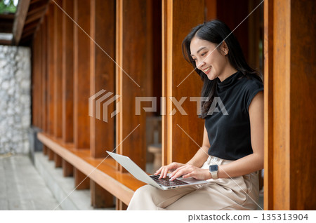 Asian woman student looking and using laptop while sitting on wooden bench in corridor or hallway of an outdoor cafe. Asian woman student looking and using laptop while sitting on wooden bench in corridor or hallway of an outdoor cafe. 135313904
