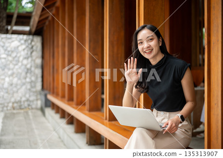 Asian woman holding laptop and waving a hand while sitting on wooden bench in a corridor or hallway of outdoor cafe Asian woman holding laptop and waving a hand while sitting on wooden bench in a corridor or hallway of outdoor cafe 135313907