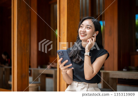 Asian woman touching earphone listening to music or on a call while sits in wooden corridor or hallway of outdoor cafe 135313909