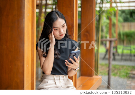 Asian woman looking at phone and touching or massaging temple while sit in cafe's wooden corridor beside bamboo garden Asian woman looking at phone and touching or massaging temple while sit in cafe's wooden corridor beside bamboo garden 135313922