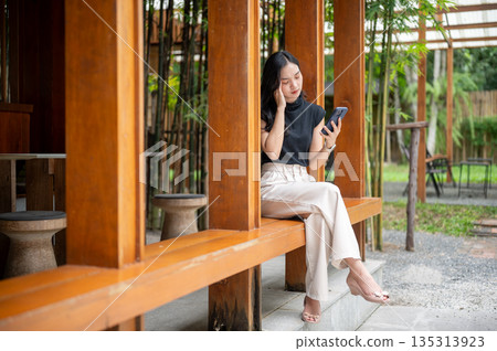 Asian woman holding phone and touching or massaging temple while sits in cafe's wooden corridor beside bamboo garden 135313923