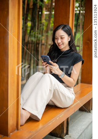 Asian woman holding phone and sitting leaning against wooden pillar in cafe's corridor beside bamboo garden 135313924