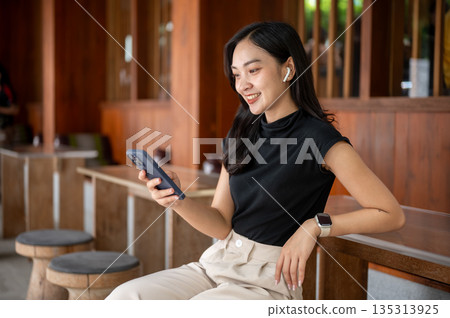 Asian woman wearings earphone is looking at phone while sitting leaning against wooden table in cafe or restaurant 135313925
