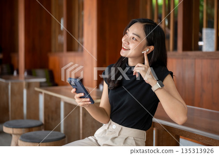 Asian woman wearings earphone is looking away while sitting leaning against wooden table in cafe or restaurant Asian woman wearings earphone is looking away while sitting leaning against wooden table in cafe or restaurant 135313926