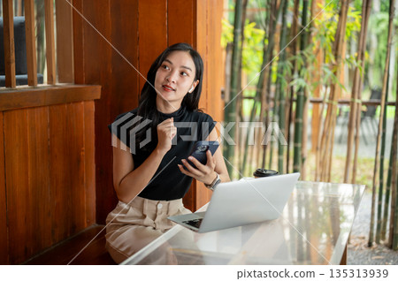 Asian woman holding and looking away from phone while sits in front of a laptop in cafe or restaurant Asian woman holding and looking away from phone while sits in front of a laptop in cafe or restaurant 135313939