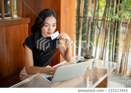 Asian woman holding credit card and looking at laptop while sitting at glass top table in the cafe. Asian woman holding credit card and looking at laptop while sitting at glass top table in the cafe. 135313942