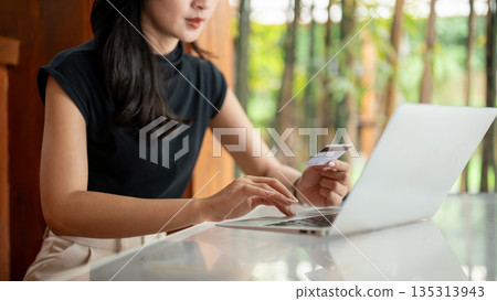 Asian woman holding a credit card and looking at laptop while sitting at glass top table in the cafe. 135313943