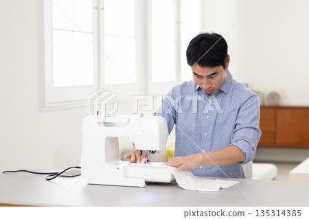 A middle-aged man sewing at a table in a bright living room A middle-aged man sewing at a table in a bright living room 135314385