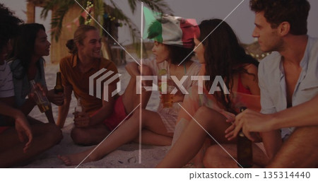 Laughing friends sitting around beach fire at dusk, holding bottles under palm trees 135314440