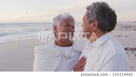 Laughing senior couple sharing laugh on sandy beach, with white pom-pom shawl and collared shirt Laughing senior couple sharing laugh on sandy beach, with white pom-pom shawl and collared shirt 135314482