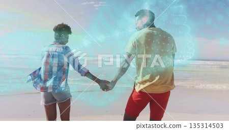 Walking couple holding hands along sandy beach, wearing yellow T-shirt, with holographic data swirl 135314503