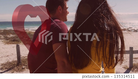 Couple in tshirt, sweatshirt gazing at sea from beach railing posts, with translucent heart graphic 135314511