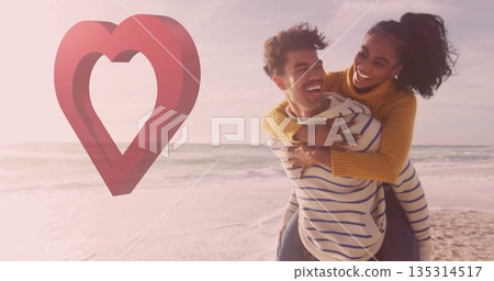 Laughing man carrying woman piggyback on sandy shoreline with waves, heart overlay, copy space 135314517