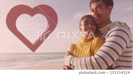 Embracing couple wearing mustard navy-striped sweaters on sandy beach, with ocean waves, heart icon 135314524