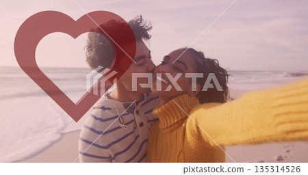 Couple wearing sweater and hoodie capturing selfie, kissing on beach, with smartphone heart overlay Couple wearing sweater and hoodie capturing selfie, kissing on beach, with smartphone heart overlay 135314526