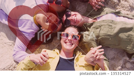 Smiling group of four women lying in circle on sandy beach, with bright sunglasses, summer clothing 135314527