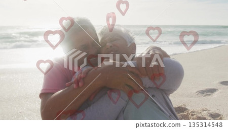 Embracing senior couple sitting on beach edge, displaying wedding rings and heart-shaped graphics 135314548
