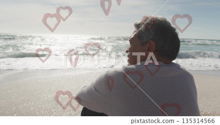 Sitting senior Hispanic man gazing at ocean waves on shoreline, with red heart overlays, copy space 135314566