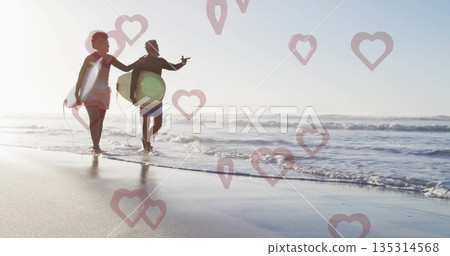 Walking couple in swimwear carrying surfboards with leashes along beach at low tide, heart graphics Walking couple in swimwear carrying surfboards with leashes along beach at low tide, heart graphics 135314568
