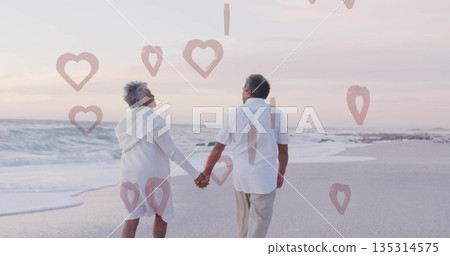 Walking senior couple holding hands along beach at dusk on wet sand, with heart-shaped graphics Walking senior couple holding hands along beach at dusk on wet sand, with heart-shaped graphics 135314575