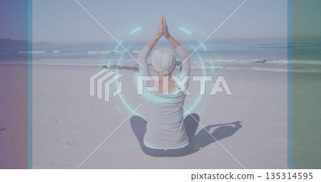 Practicing senior woman raising arms in prayer on sandy beach, with digital holographic overlay 135314595