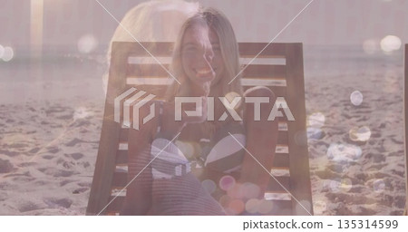 Sitting woman wearing bikini on lounge chair at beach, with lens flares and calm ocean horizon 135314599