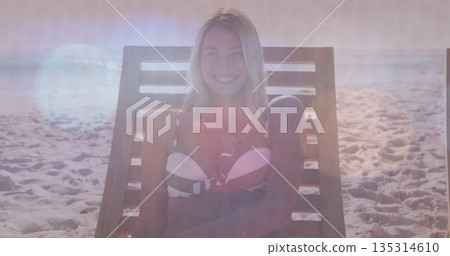 Smiling woman wearing bikini top sitting on lounge chair on beach, with ocean horizon 135314610