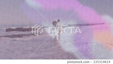 Walking couple wearing summer clothes holding arms along beach, with gentle surf and rocky outcrops 135314614