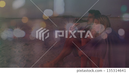 Sipping couple wrapped in fringed throw blanket on beach at dusk, with glass bottles, copy space 135314641