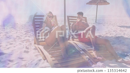 Reclining couple wearing swimwear and sunglasses on tropical beach, with wooden chairs, cold drinks 135314642