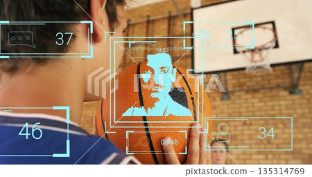Raising basketball close to cheek, teenage boy wearing jersey in school gym with overlay graphics Raising basketball close to cheek, teenage boy wearing jersey in school gym with overlay graphics 135314769