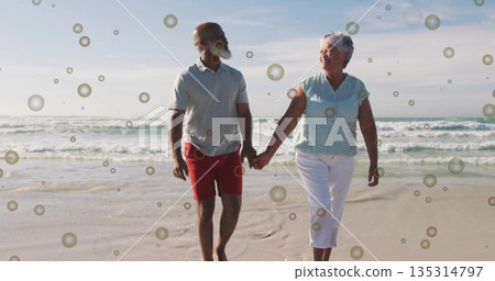 Walking couple holding hands in grey polo, red shorts, blue top on ocean sand, with bubbles 135314797