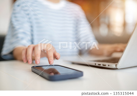Close up of young woman hands touching on phone and typing on laptop while sitting at table in cafe Close up of young woman hands touching on phone and typing on laptop while sitting at table in cafe 135315044