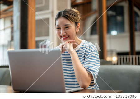 Asian young adult woman clasping hand under chin looking at laptop while sit at wooden table in cafe Asian young adult woman clasping hand under chin looking at laptop while sit at wooden table in cafe 135315096