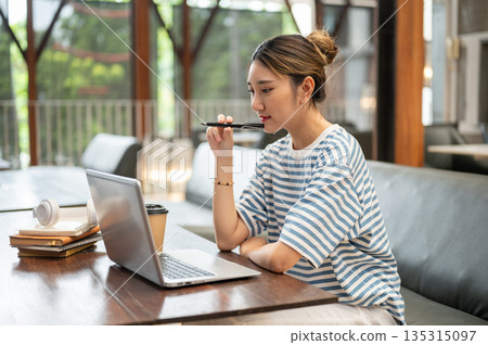 Asian young adult woman holding fountain pen and looking at laptop while sit at wooden table in cafe Asian young adult woman holding fountain pen and looking at laptop while sit at wooden table in cafe 135315097