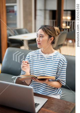 Asian young woman holding pen looking away from book while sits on sofa across laptop at table in cafe. Asian young woman holding pen looking away from book while sits on sofa across laptop at table in cafe. 135315102