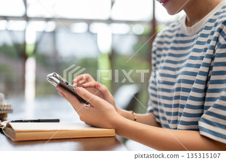 Close up of a woman holding and looking at smartphone over book while sitting at wooden table in cafe. Close up of a woman holding and looking at smartphone over book while sitting at wooden table in cafe. 135315107