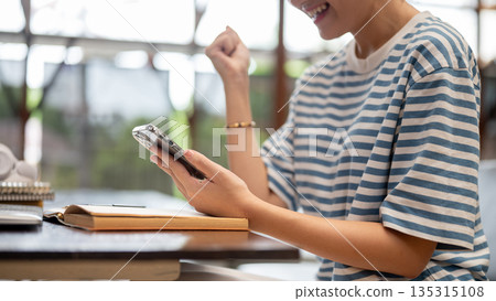 Close up of a woman holding phone looking excited over a book while sits at wooden table in the cafe 135315108