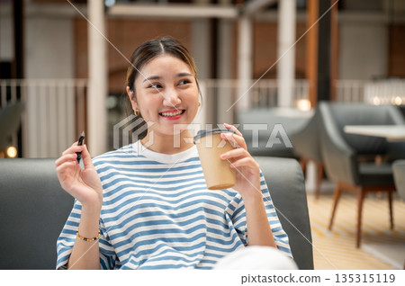 Smiling asian young woman holding pen and coffee cup as she looking around while sit on sofa in cafe Smiling asian young woman holding pen and coffee cup as she looking around while sit on sofa in cafe 135315119