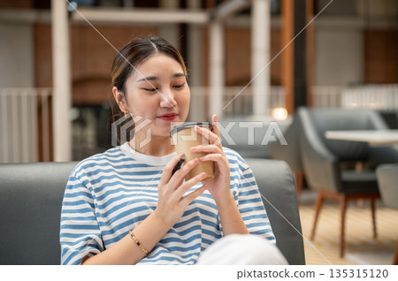 Asian young woman holding and smelling coffee aroma inside a cup while sitting on sofa in the cafe. Asian young woman holding and smelling coffee aroma inside a cup while sitting on sofa in the cafe. 135315120