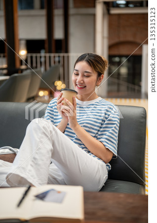 Young asian woman looking at coffee holding with both hands while sitting leaning on sofa in a cafe. Young asian woman looking at coffee holding with both hands while sitting leaning on sofa in a cafe. 135315123