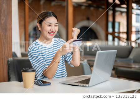 Happy asian woman holding credit card while sitting with laptop at table in a cafe or coffee shop. 135315125