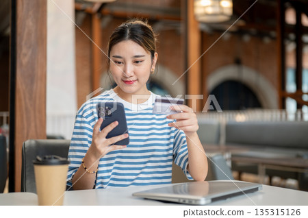 Asian woman holding credit card and looking at phone while sitting at table in cafe or coffee shop. 135315126