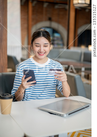 Smiling asian woman holding credit card looking at phone while sits at table in cafe or coffee shop. 135315127