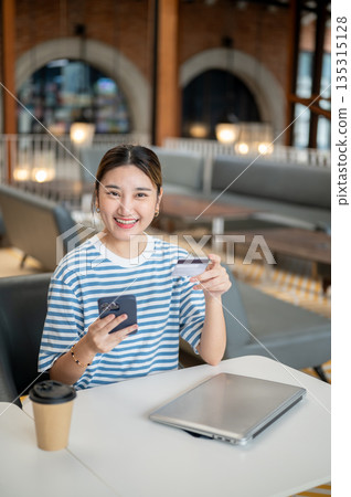 Smiling asian woman holding credit card and smartphone while sits at table in cafe or coffee shop. Smiling asian woman holding credit card and smartphone while sits at table in cafe or coffee shop. 135315128