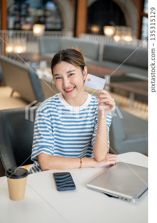 Smiling young asian woman holding or showing credit card while sits with phone and laptop in a cafe. Smiling young asian woman holding or showing credit card while sits with phone and laptop in a cafe. 135315129