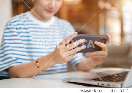 Close up of asian woman holding smartphone horizontal over laptop while sits at white table in cafe. 135315135