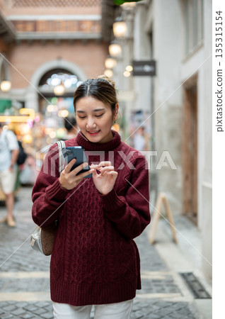 Young asian woman in knit sweater looking typing on phone while walking in shopping mall or market. Young asian woman in knit sweater looking typing on phone while walking in shopping mall or market. 135315154