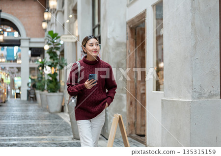 Pretty asian woman in knit sweater holding phone walking and looking around shopping mall or market. Pretty asian woman in knit sweater holding phone walking and looking around shopping mall or market. 135315159