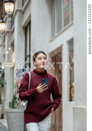 Pretty asian woman in knit sweater holding phone while walking in a shopping mall or market hall. Pretty asian woman in knit sweater holding phone while walking in a shopping mall or market hall. 135315160